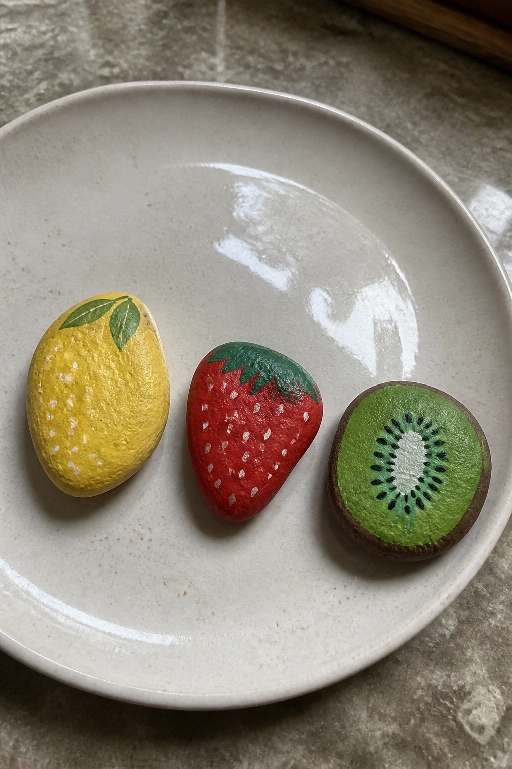 Three painted rocks resembling lemon, strawberry, and kiwi on a white plate.