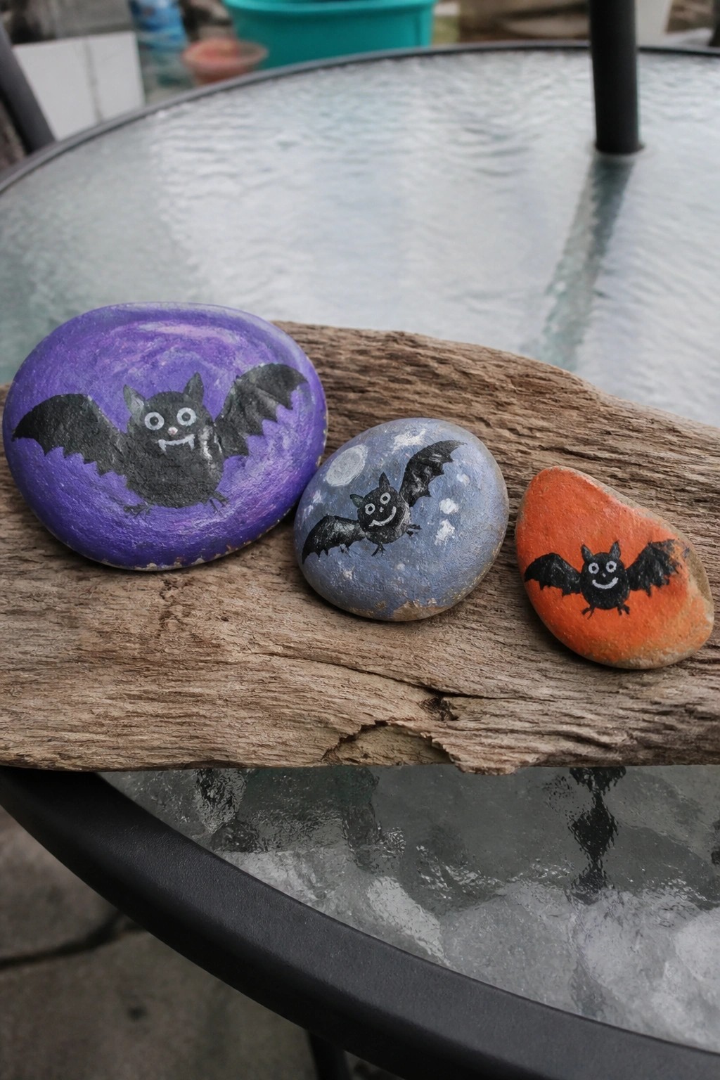 Three bat-painted rocks in purple, blue, and orange on weathered wood over glass.