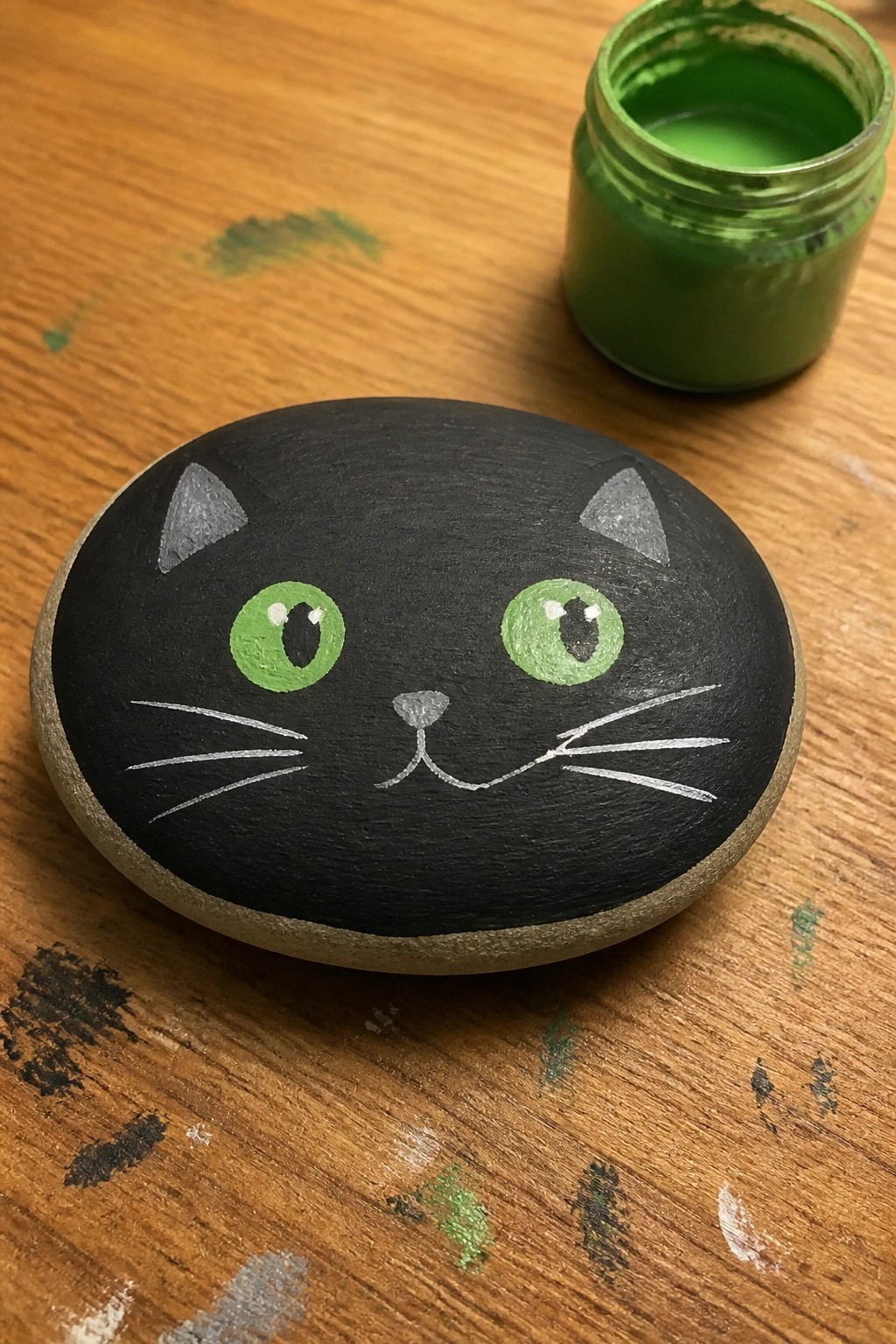 Black cat face painted on rock with green eyes beside open green paint jar on wood