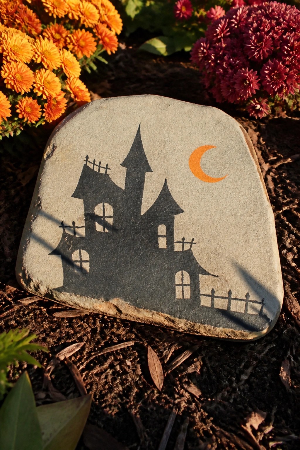 Gray stone painted with black haunted house silhouette and orange crescent moon among flowers.