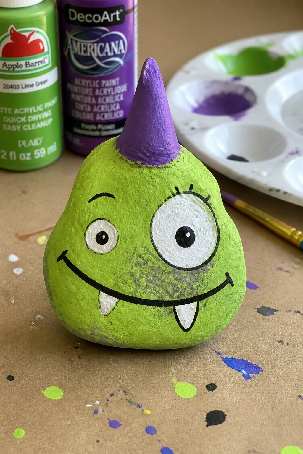 Green painted rock monster with purple horn, cartoon eyes, and fangs beside paints and palette