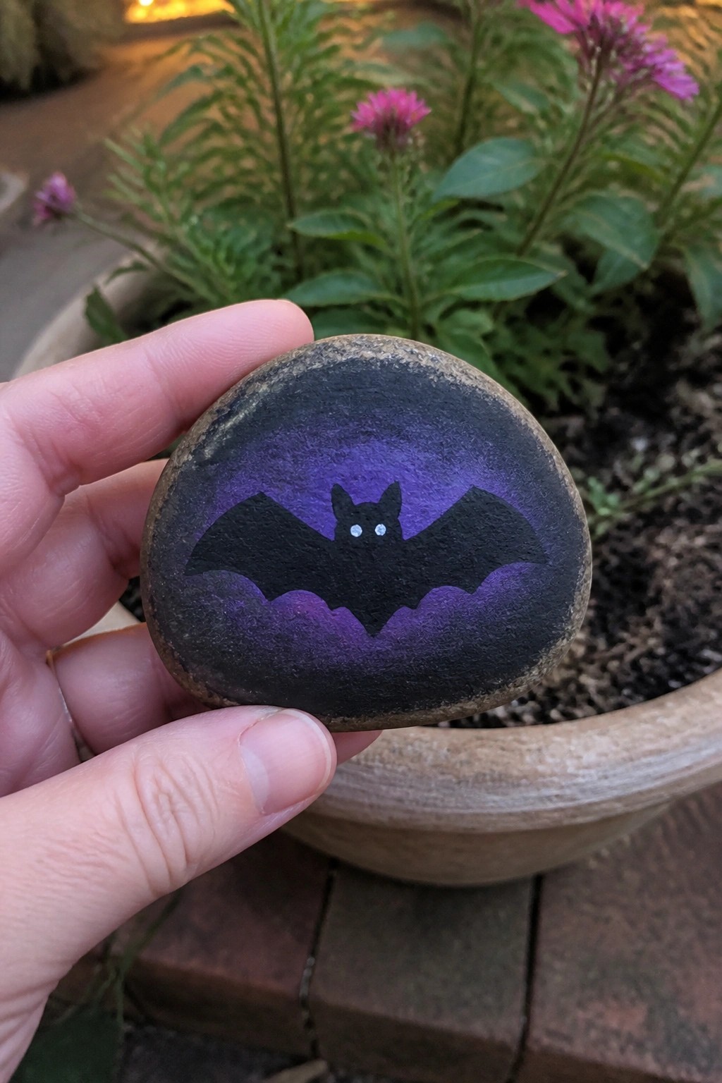 Hand holds round rock painted with black bat on purple gradient background