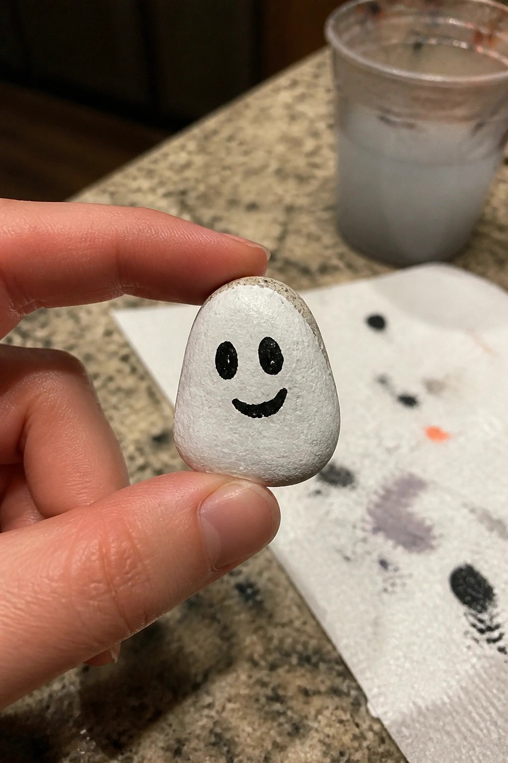 Hand holding small white rock painted with black smiley face on counter