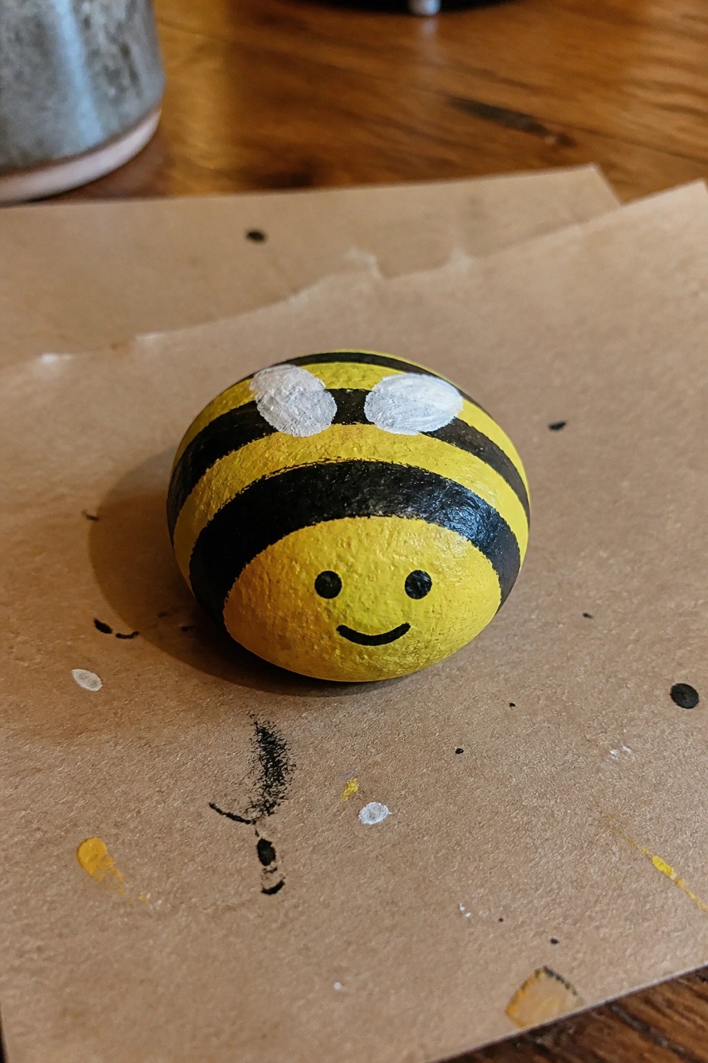 Yellow and black striped rock painted as smiling bee with white wings.