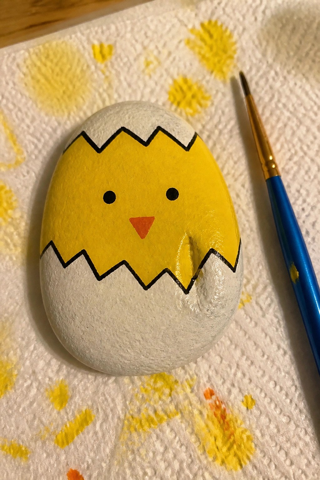 A smooth oval rock painted as a yellow chick with black eyes and an orange beak hatching from a white eggshell marked by black zigzag cracks, resting on a paper towel beside a blue paintbrush.