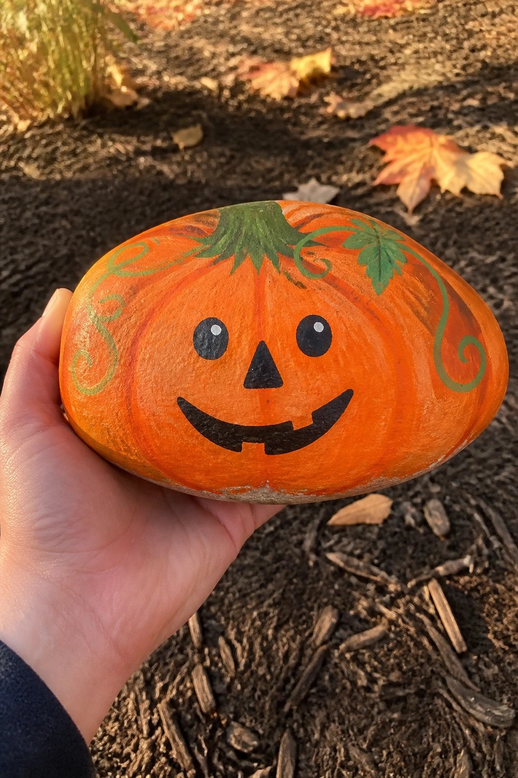 Hand holding orange painted rock pumpkin with black smiling jack-o'-lantern face and green vines.