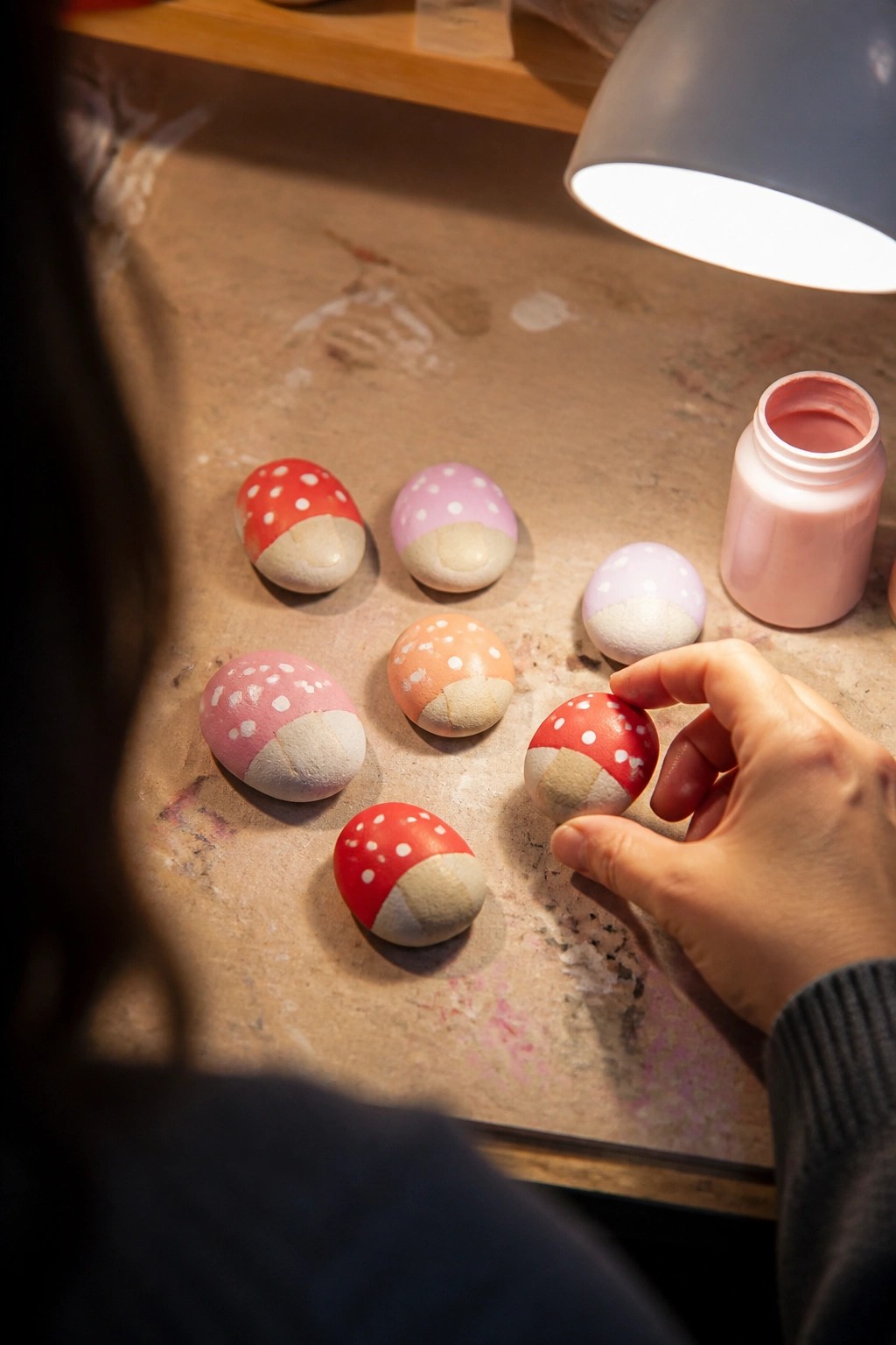 Hand holding red mushroom-painted egg among colorful ones on splattered table.