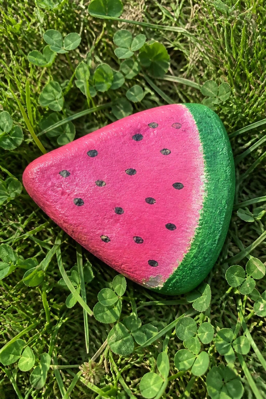 Watermelon-slice painted rock resting on green grass with clover leaves