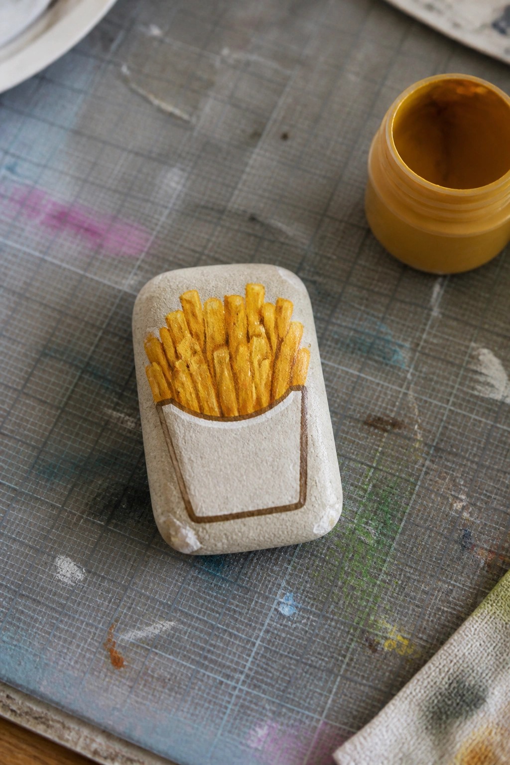 Painted rock with golden French fries in a brown carton on a gridded mat.