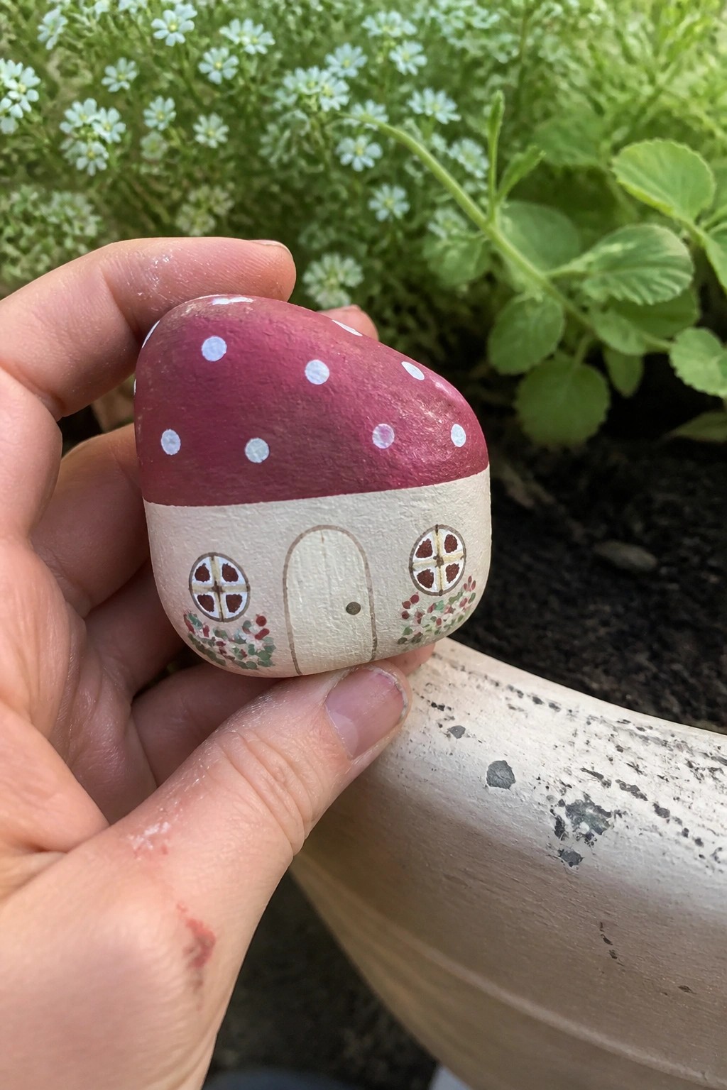 A hand holds a small painted rock shaped like a red mushroom house with white dots, a door, windows, and flowers.