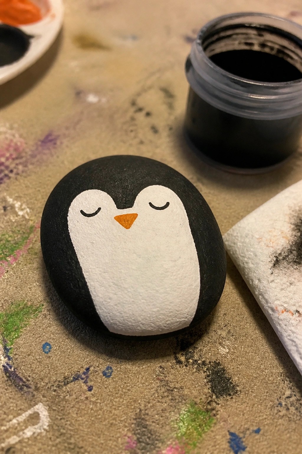 Painted rock penguin with closed eyes and orange beak on paint-splattered table