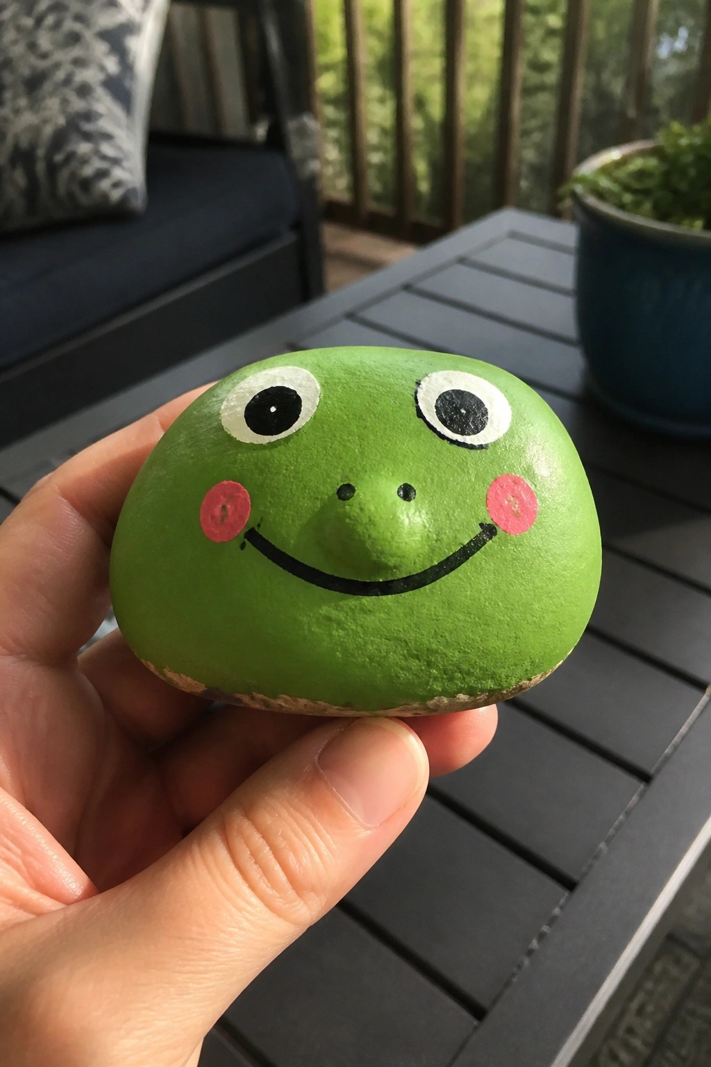 Hand holding a green rock painted as a smiling frog with pink cheeks