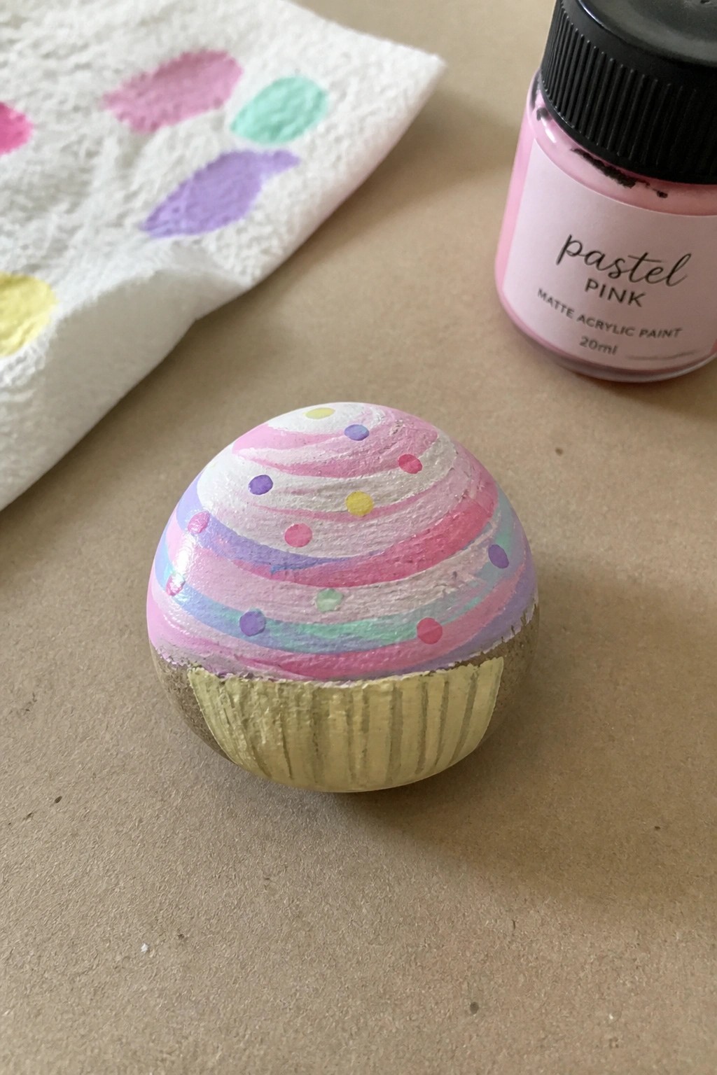 A small rounded rock painted to look like a cupcake with a gold base and pastel swirled frosting plus colorful dots.
