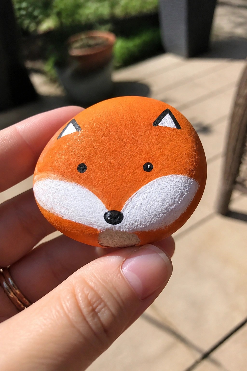 A hand holds a small round orange rock painted as a fox face with white muzzle and black eye and nose dots.