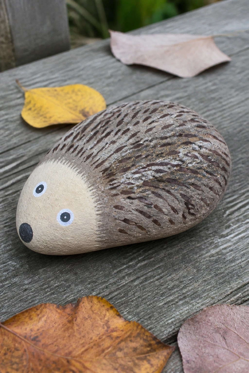 Painted hedgehog stone on wooden surface amid scattered autumn leaves