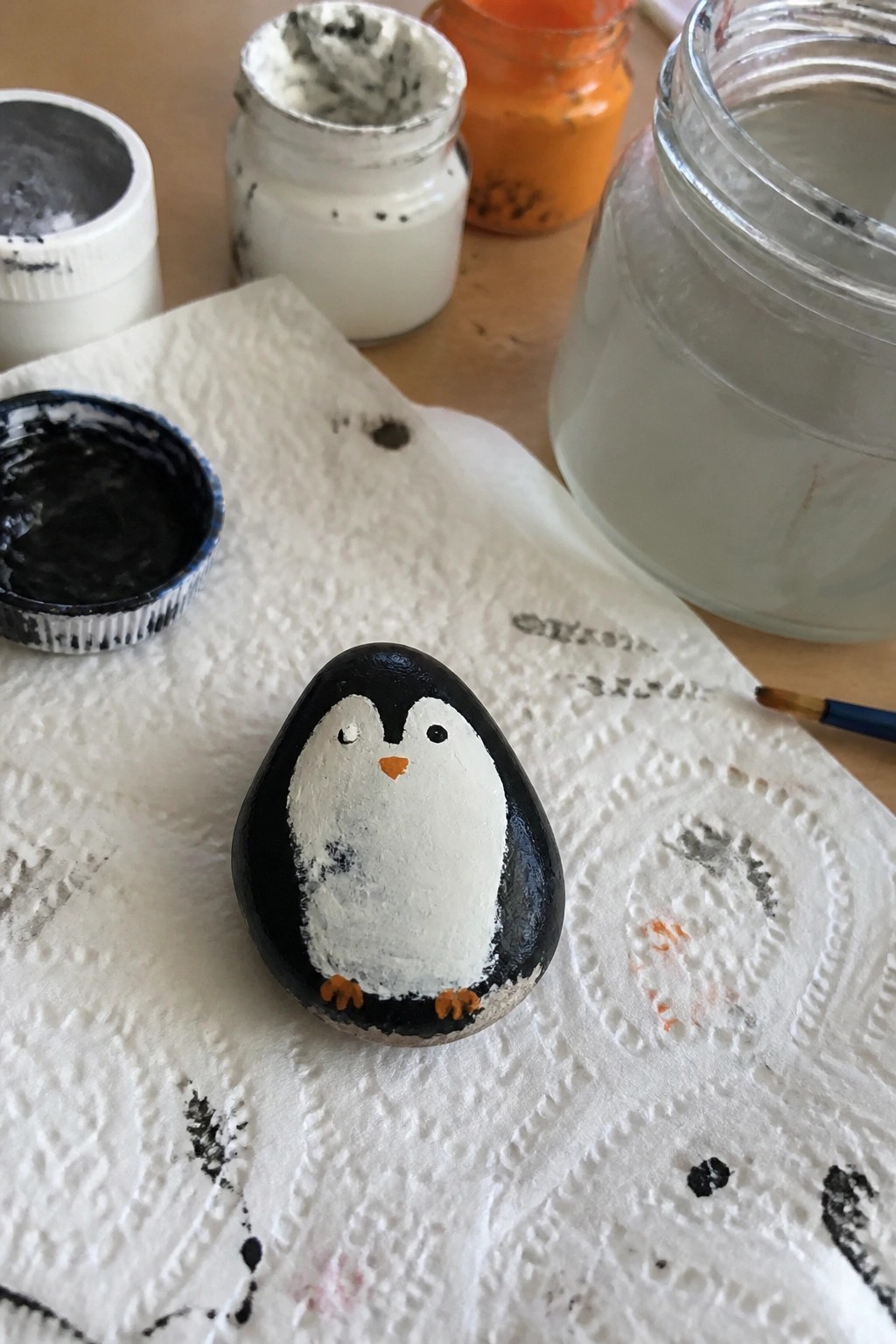 A small black and white penguin painted on an egg-shaped rock sits on a paper towel surrounded by jars of paint.