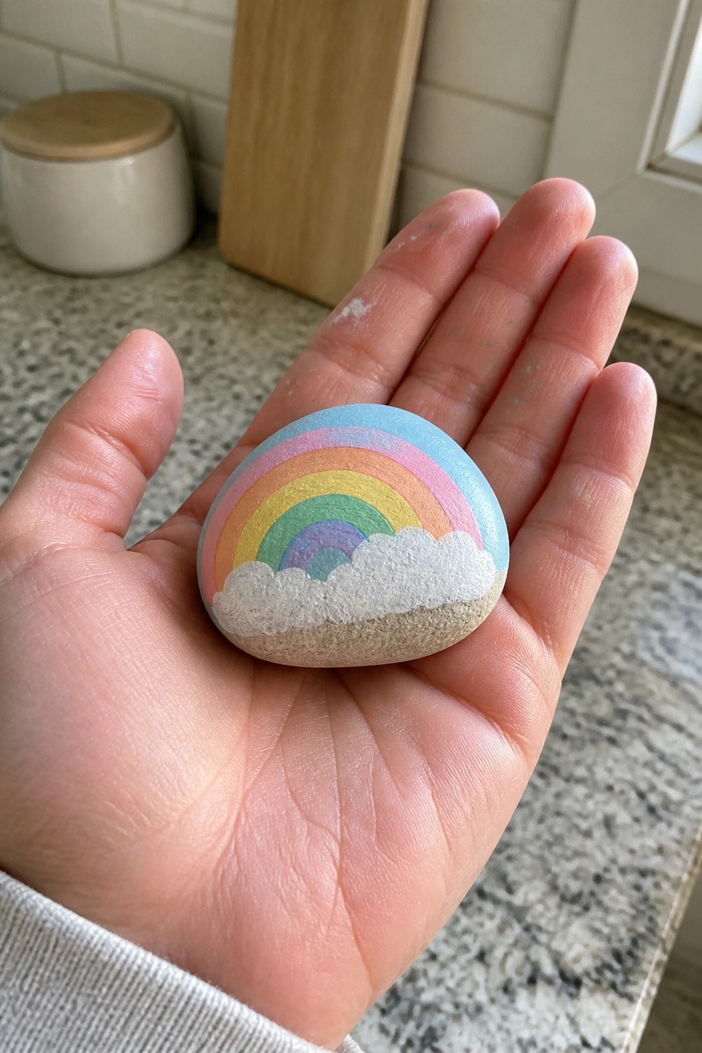 Hand holding painted stone with colorful rainbow arching over white clouds