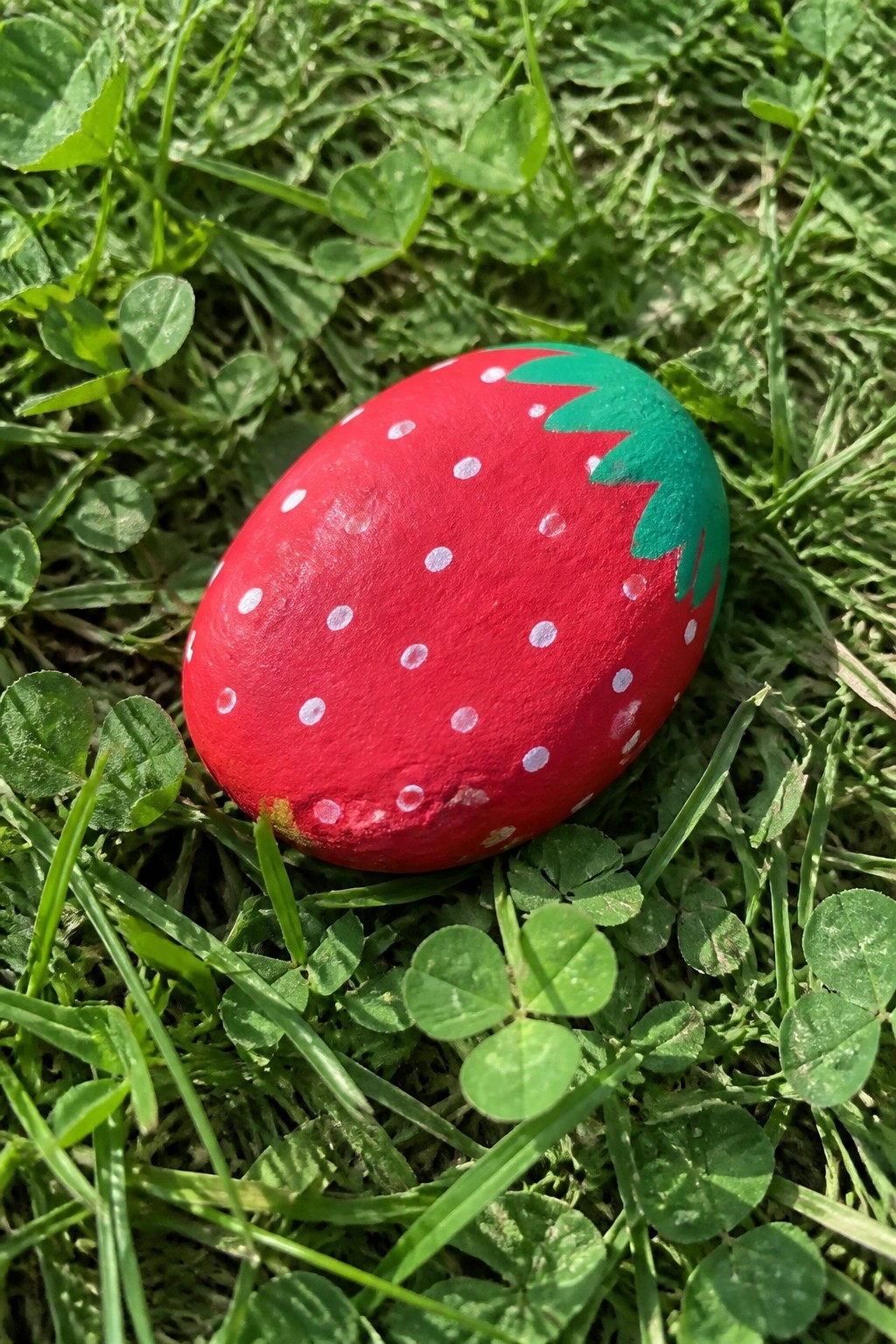 Red painted rock resembling a strawberry with white dots and green top on grass.