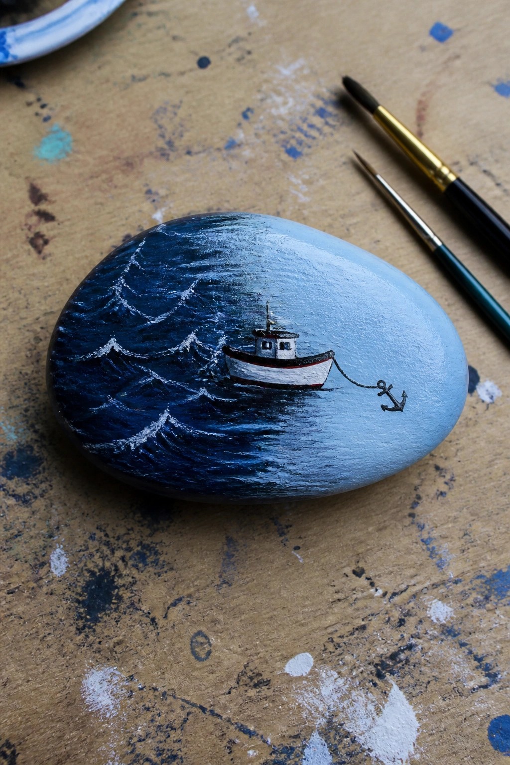 Painted stone with boat on stormy blue waves, anchor chain, and paintbrushes nearby.