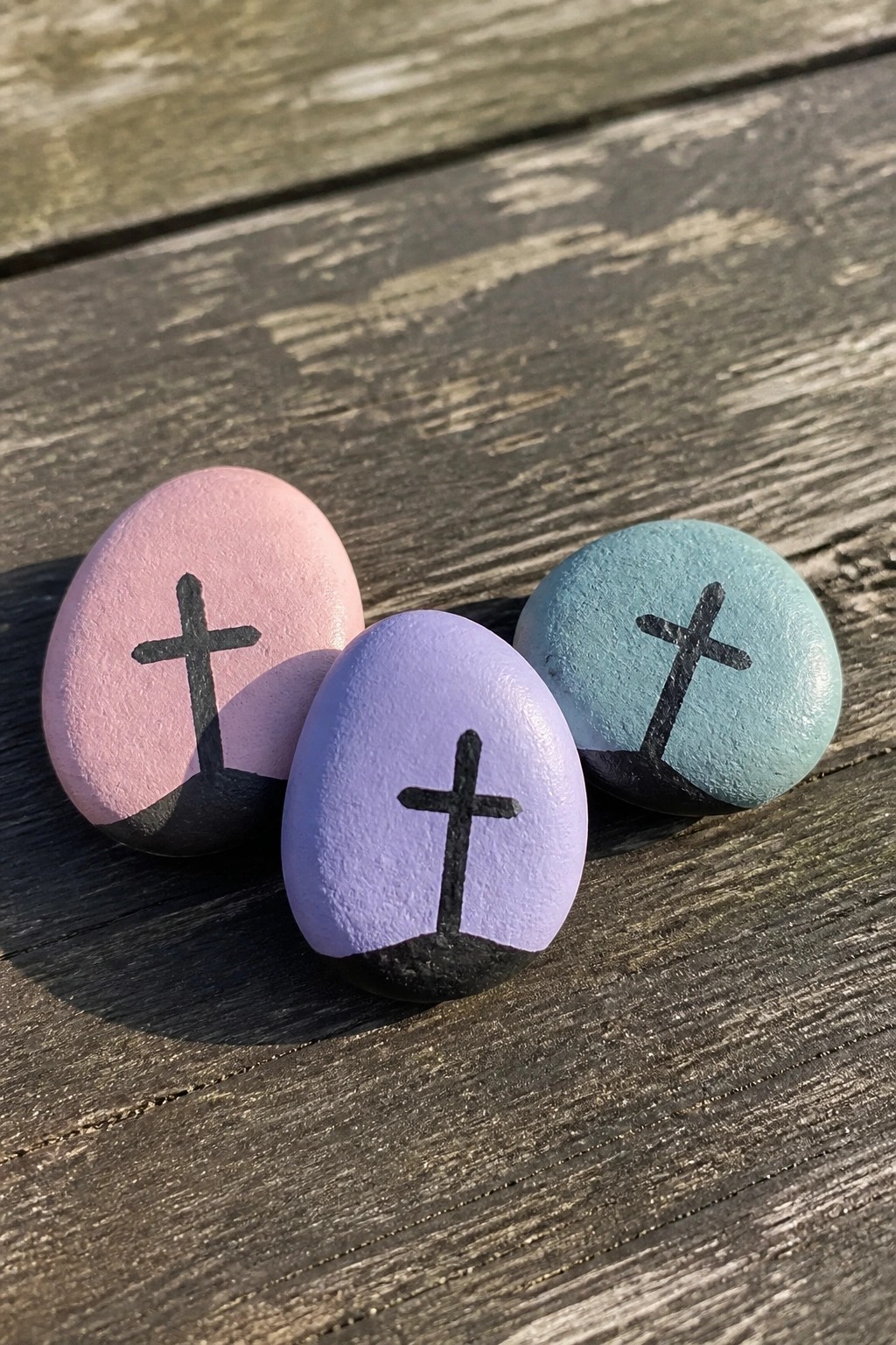 Three smooth stones painted pink, purple, and blue with black crosses rest on a wooden surface.