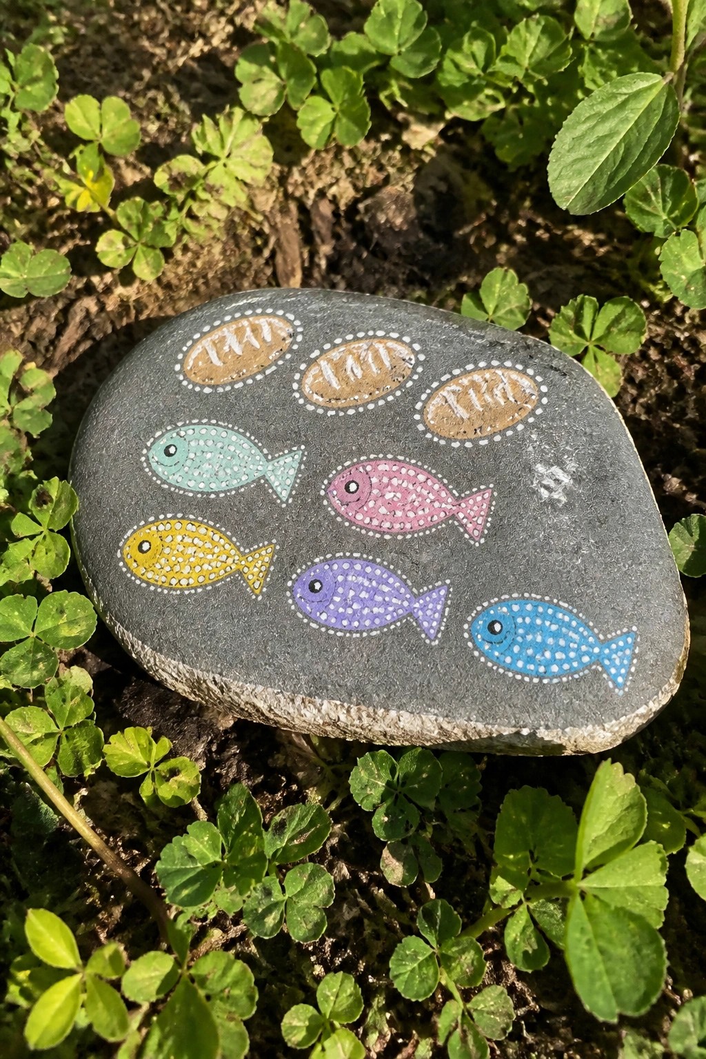 Painted gray stone with colorful dotted fish and brown ovals on green clover