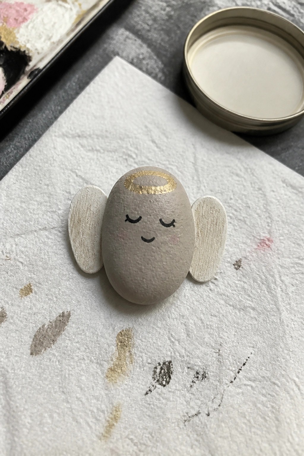 Painted stone angel with gold halo, closed eyes, pink cheeks, and white wings on paper.