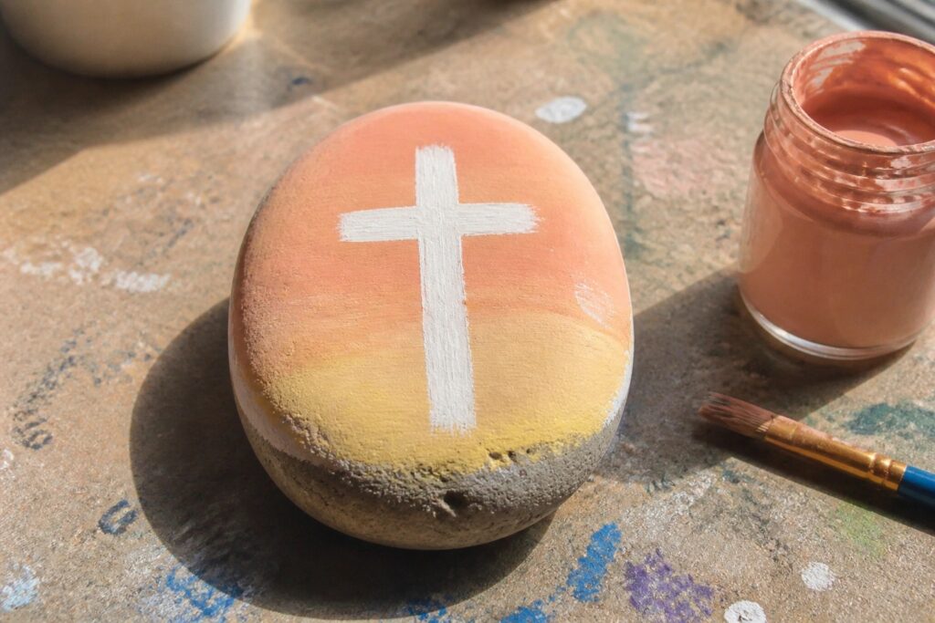 Painted rock with white cross on coral-yellow gradient beside paint jar and brush