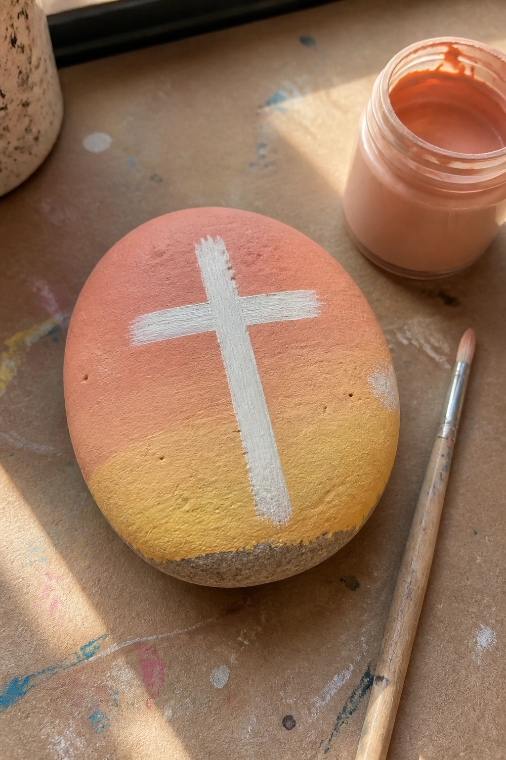Painted rock with white cross on coral-yellow gradient beside paint jar and brush