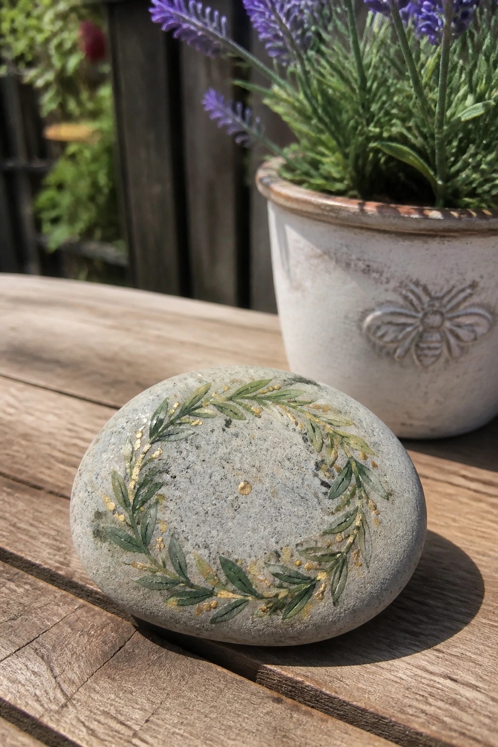 A gray rounded stone painted with a green and gold wreath sits on a wooden table next to a potted plant with purple flowers.
