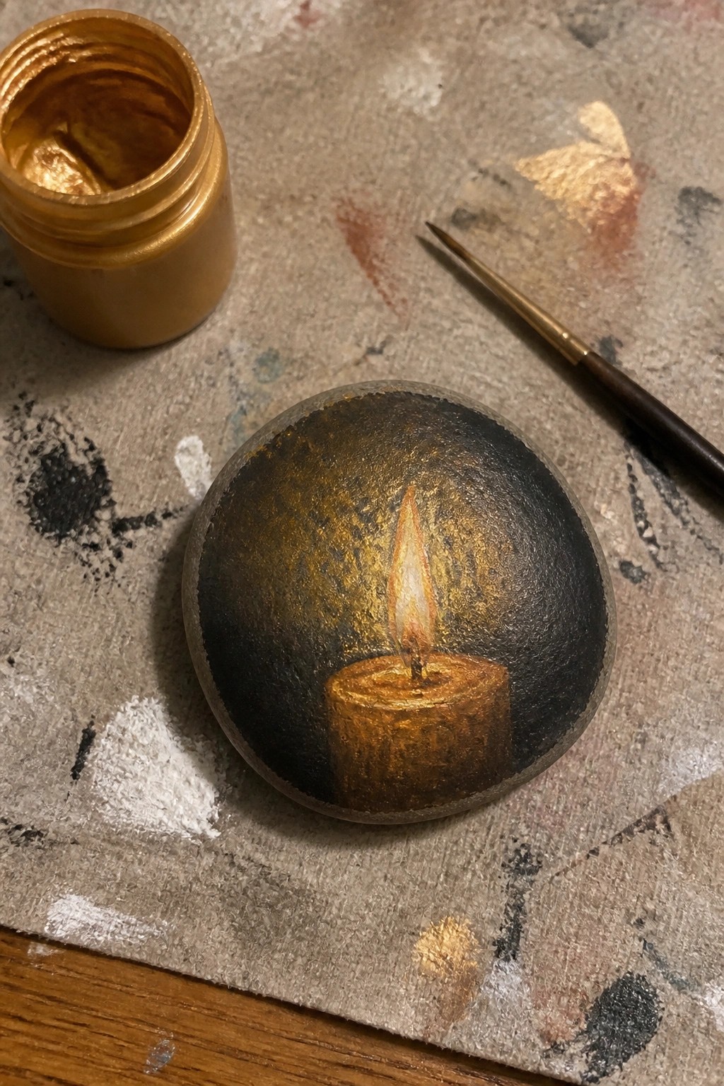 Painted stone with glowing candle beside gold paint jar and thin brush