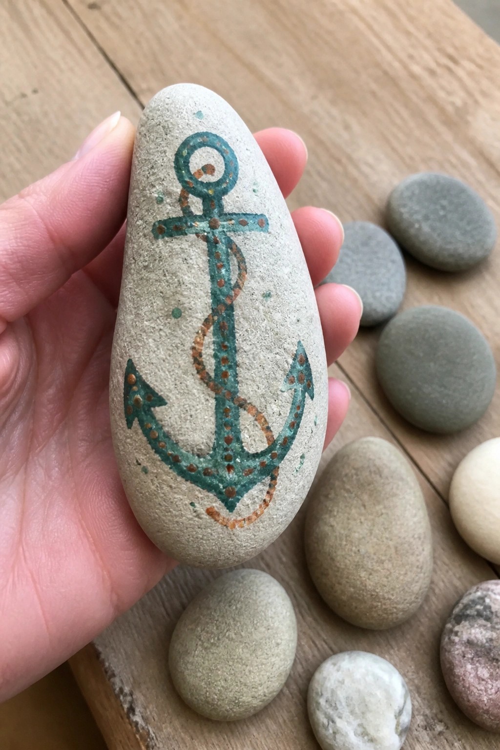 Hand holds beige stone painted with teal dotted anchor and brown rope
