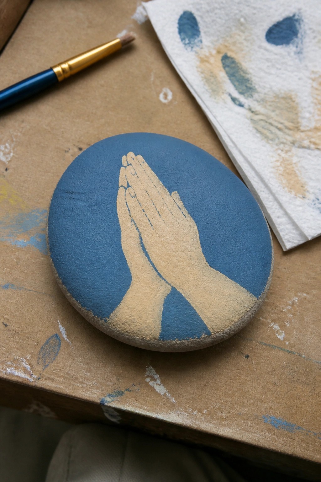 Blue painted stone with beige praying hands beside brush and stained paper towel