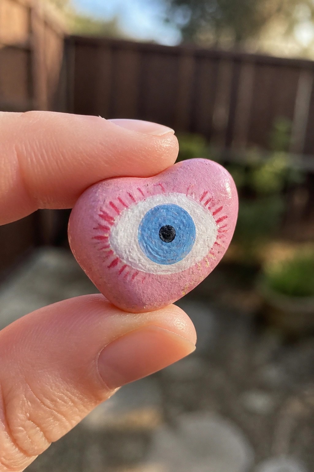A hand holds a small pink heart-shaped rock painted with a blue evil eye design that has red lashes.
