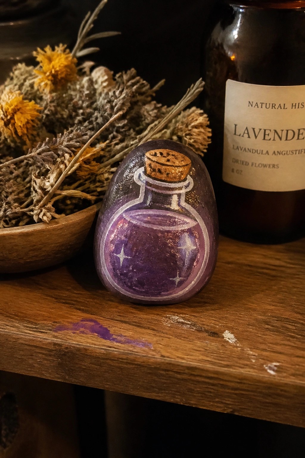 Painted rock resembling a purple potion bottle with cork on wooden table beside dried herbs