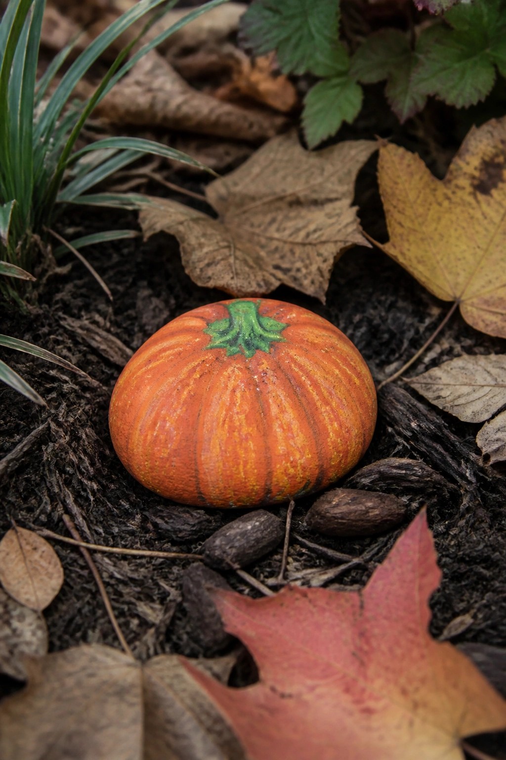 Painted orange pumpkin rock on soil amid dry autumn leaves and twigs.