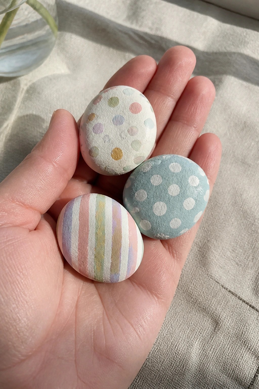 Hand holding three pastel eggs with dots and stripes on light fabric.