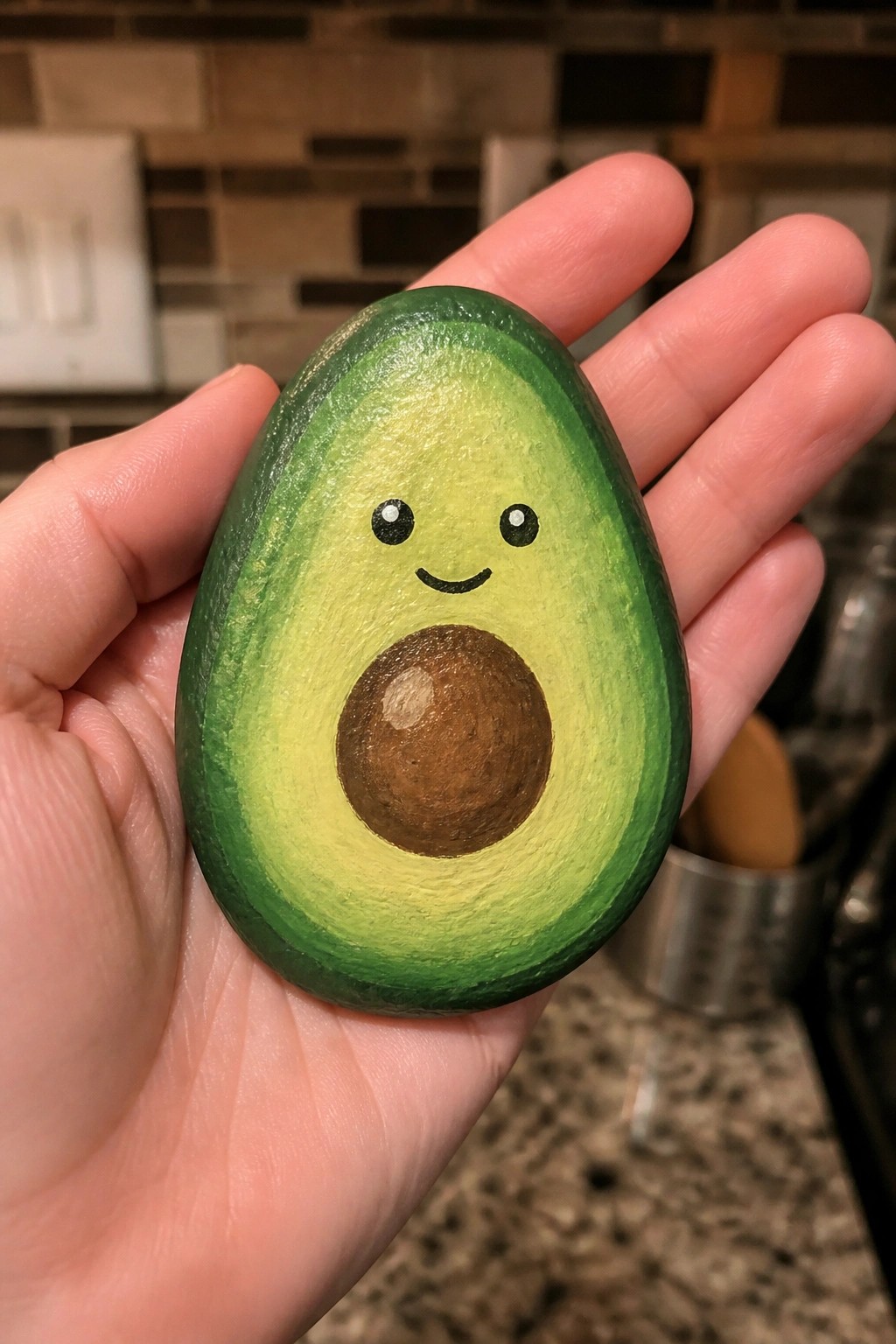 Hand holding a painted avocado rock with smiling cartoon face and pit.