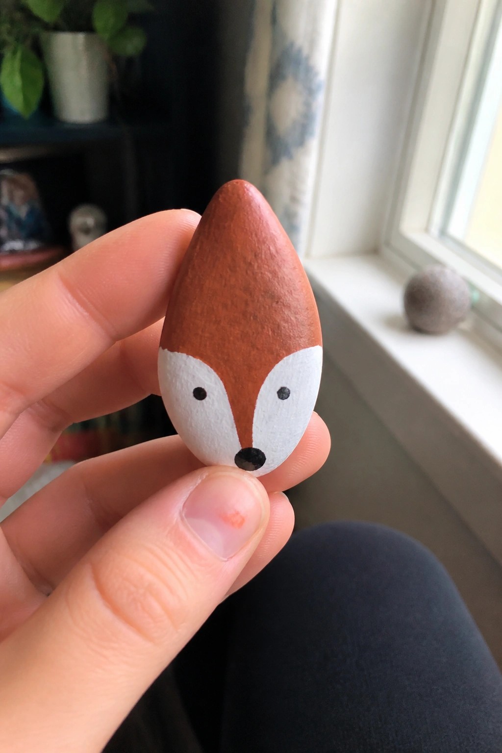 Hand holding a teardrop pebble painted as a fox with brown, white, and black.
