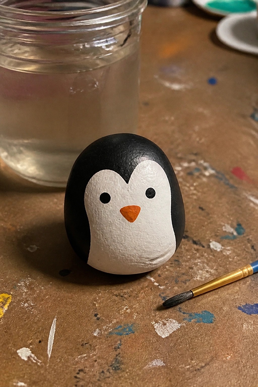Painted penguin rock with black head, white belly, orange beak on splattered table