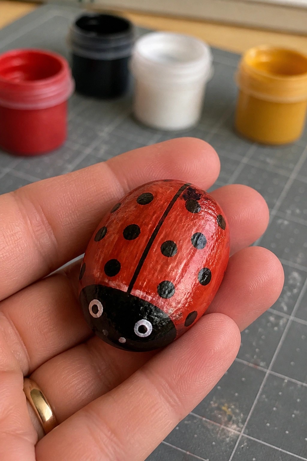 Hand holding red ladybug-painted rock with black spots near paint pots