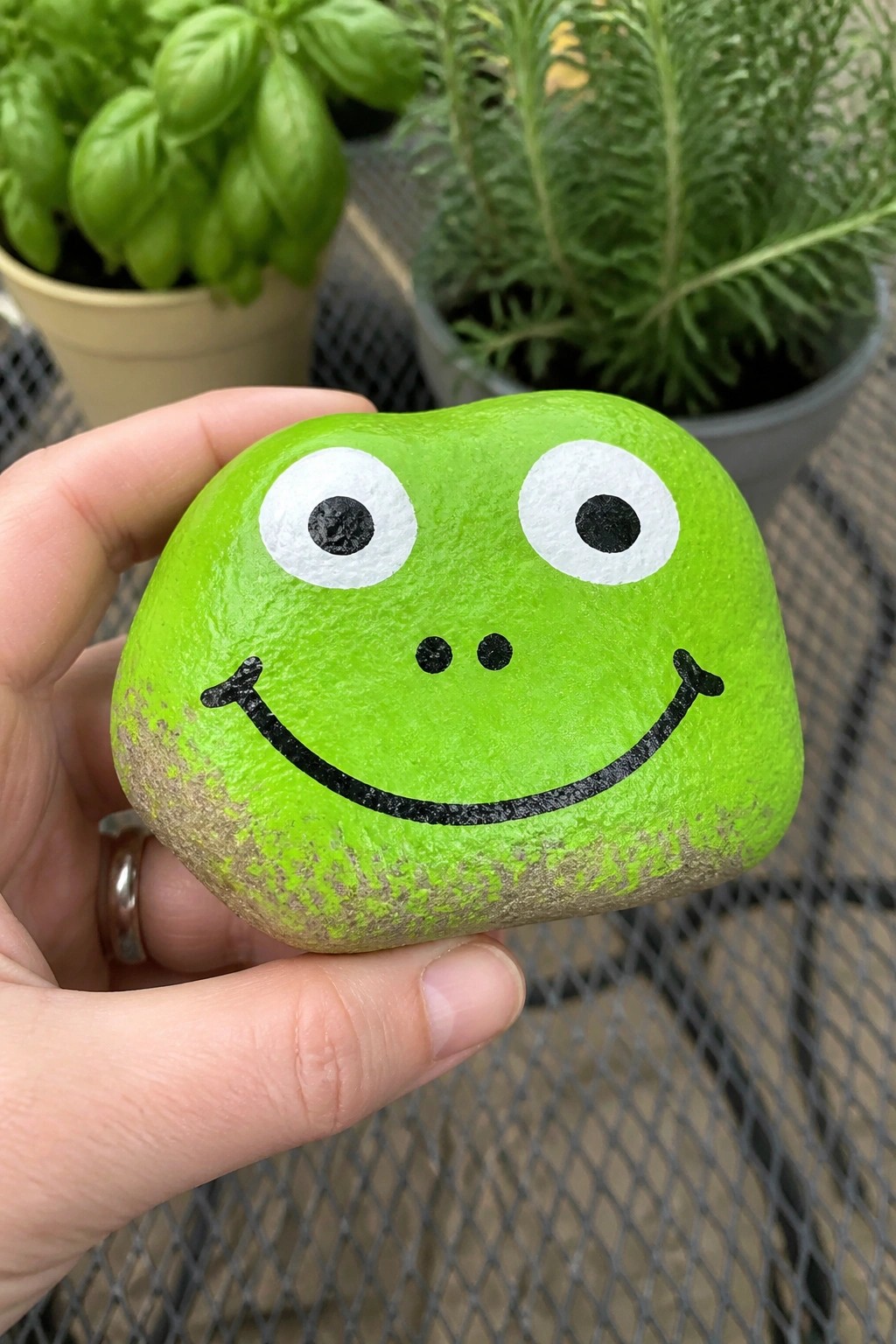 Hand holding bright green rock painted as smiling frog with white eyes