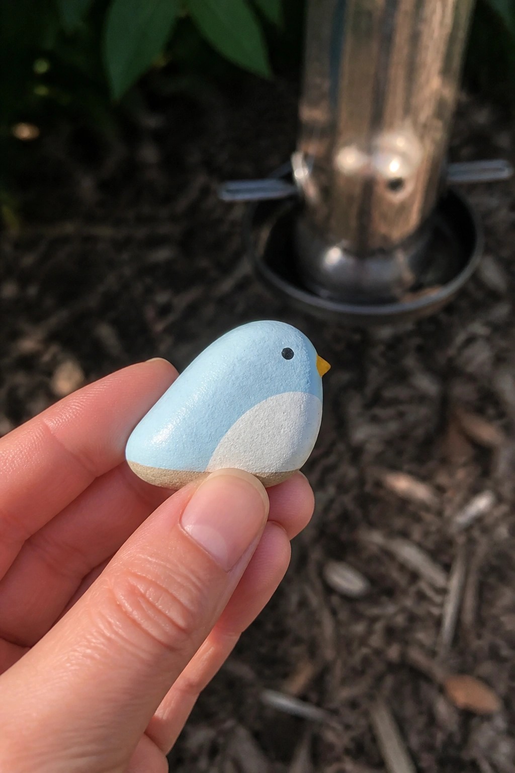 Hand holding small light blue painted bird pebble with white belly and yellow beak