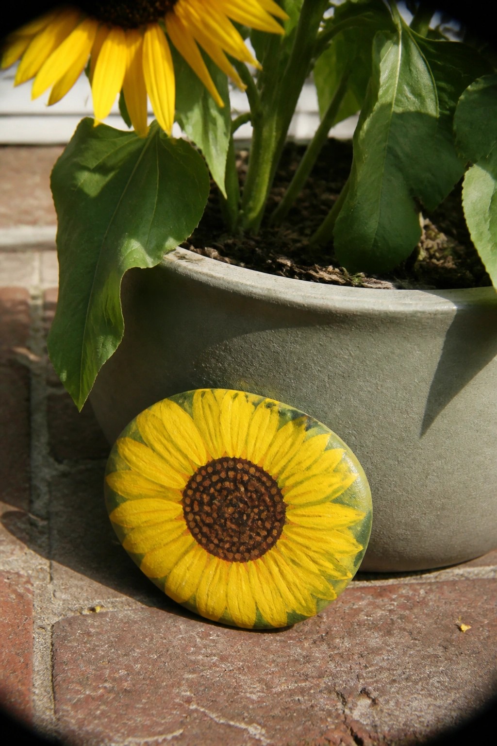 Painted sunflower rock on bricks beside potted plant in gray pot