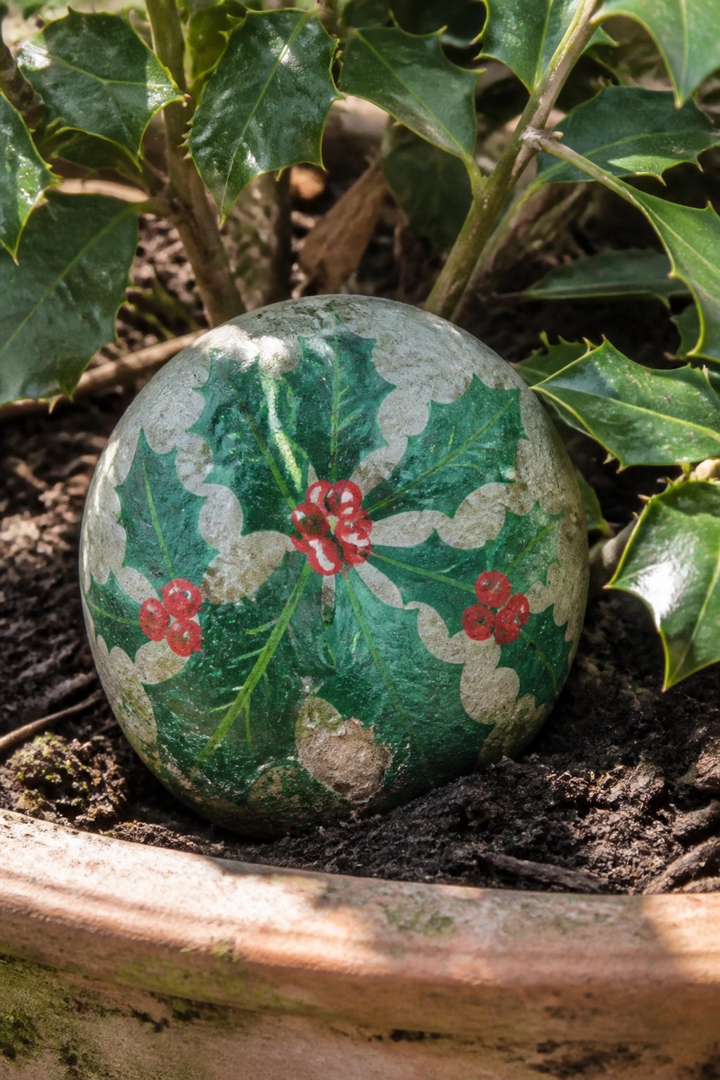 Painted stone with green holly leaves and red berries on soil