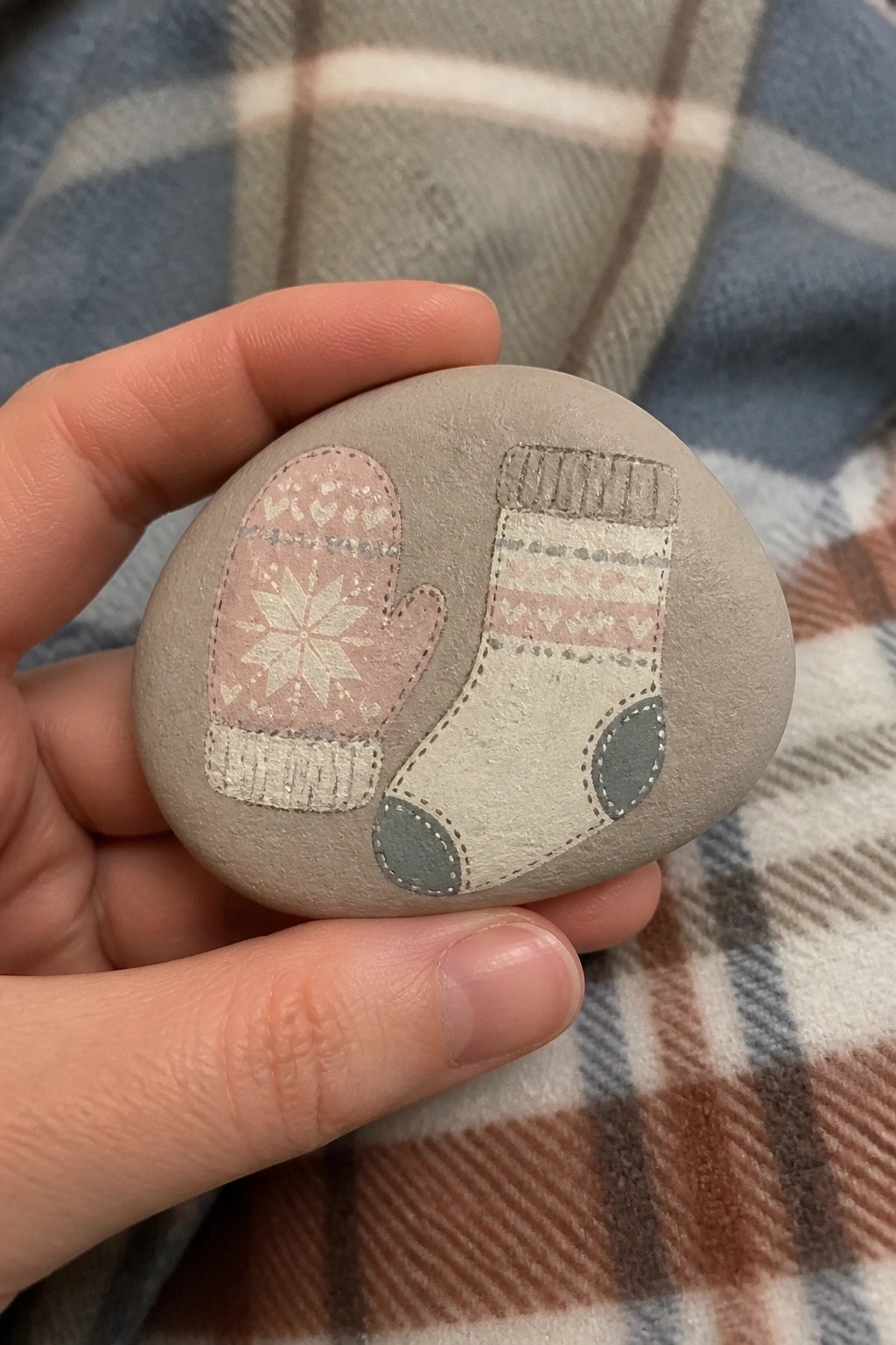 Hand holding gray stone painted with pink mitten and patterned sock on plaid fabric