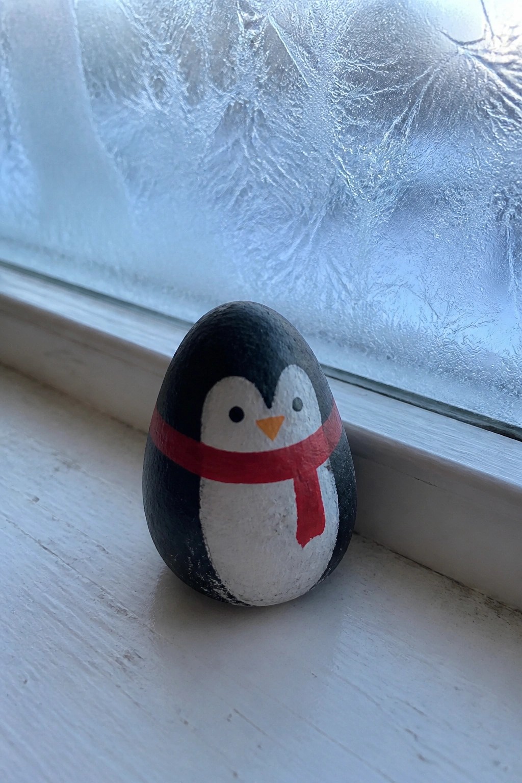 Painted black-and-white penguin rock with red scarf on frosted windowsill