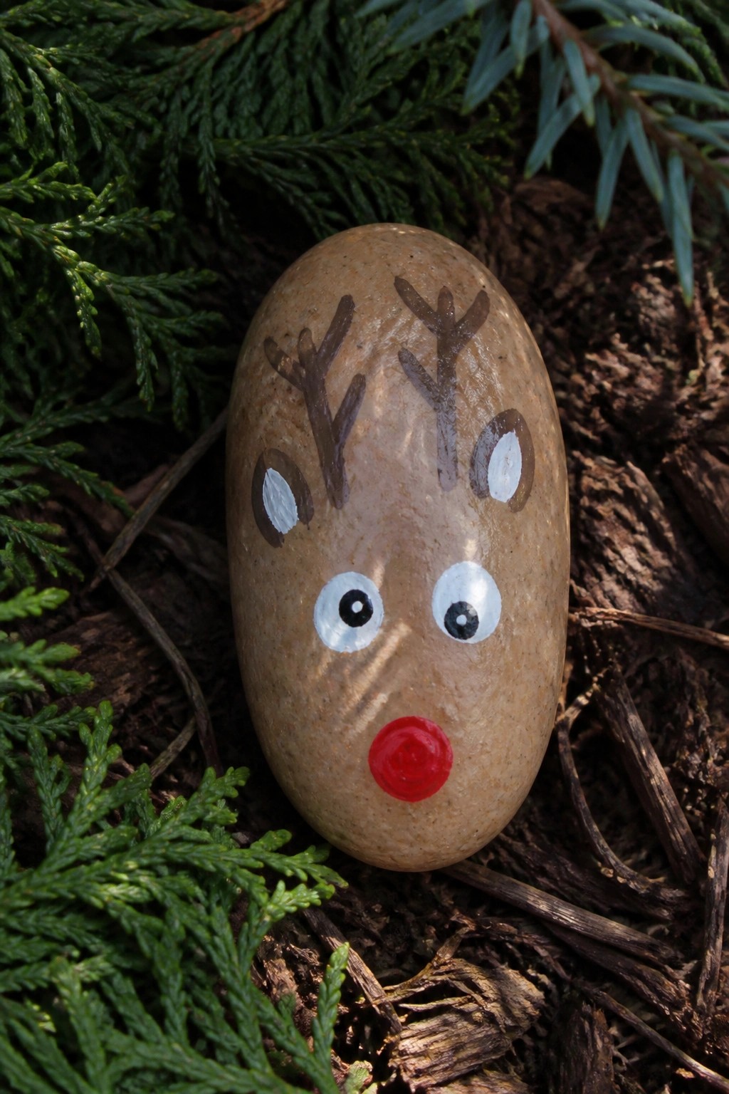 Brown rock painted as reindeer with antlers, white eyes, and red nose amid greenery.