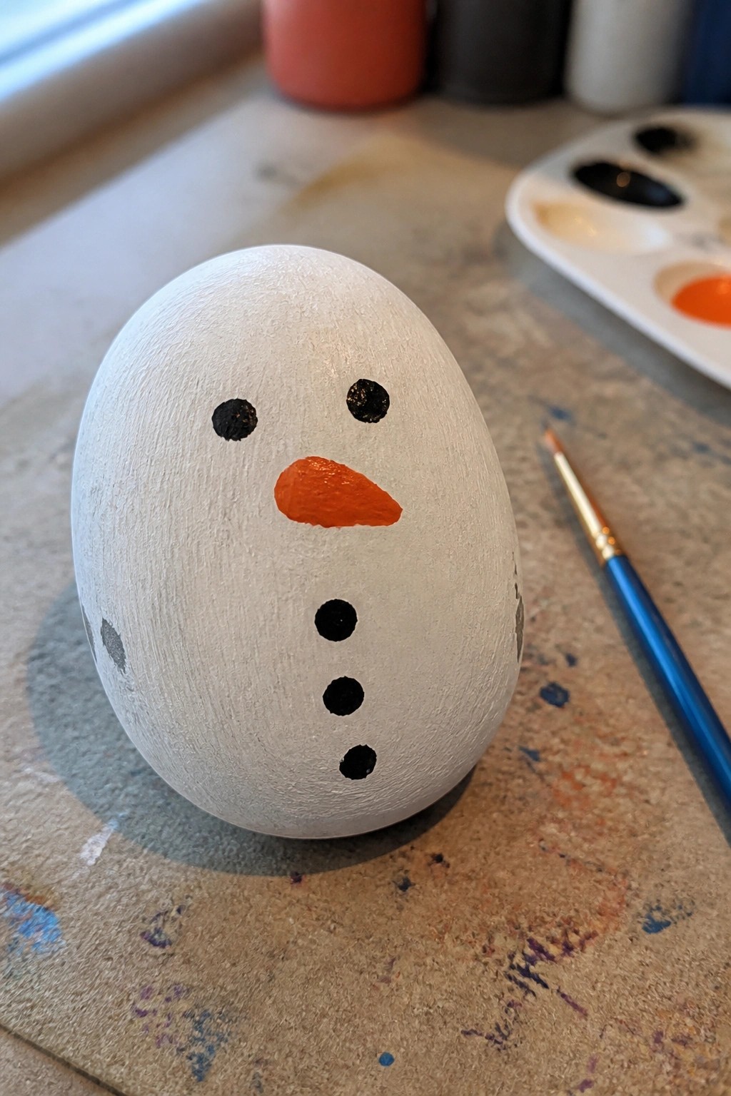 White egg snowman with black eyes, buttons, orange nose on paint-splattered table