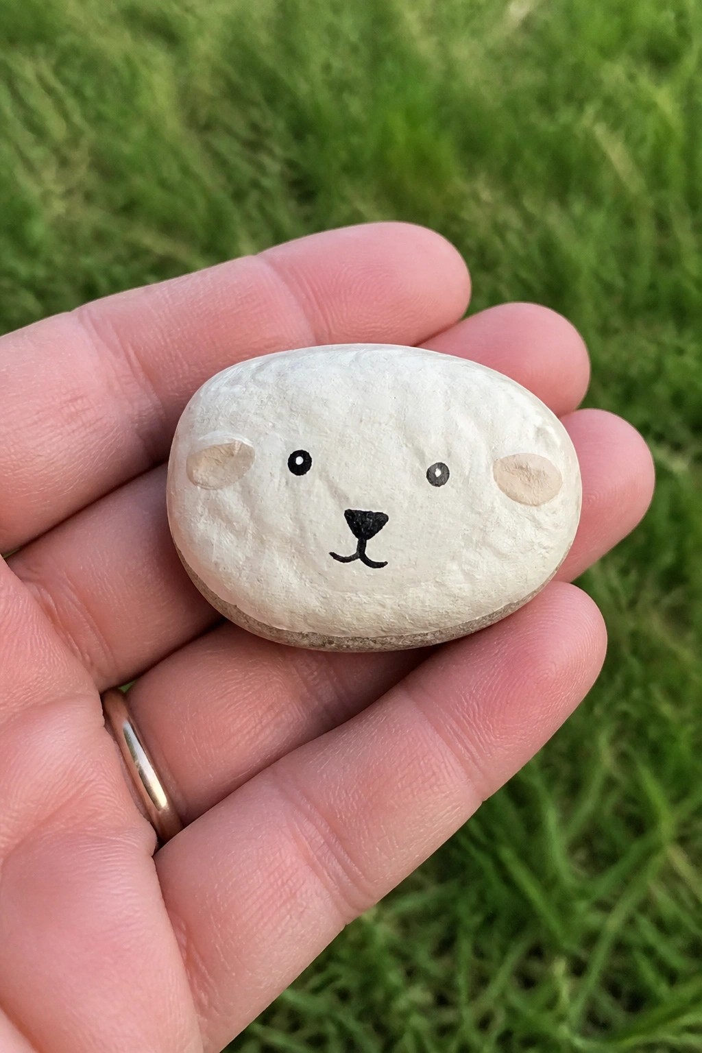 Hand holding white painted stone bear face with black features on green grass.