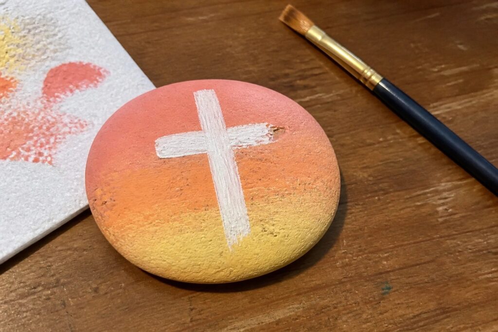 Oval rock with orange-yellow gradient and white cross beside paintbrush on wooden table